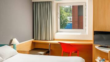 A hotel room with a bed, a desk, a red chair, a television, and a window with curtains.