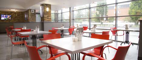 A restaurant with red chairs and white tables, large windows, and a TV on the wall.