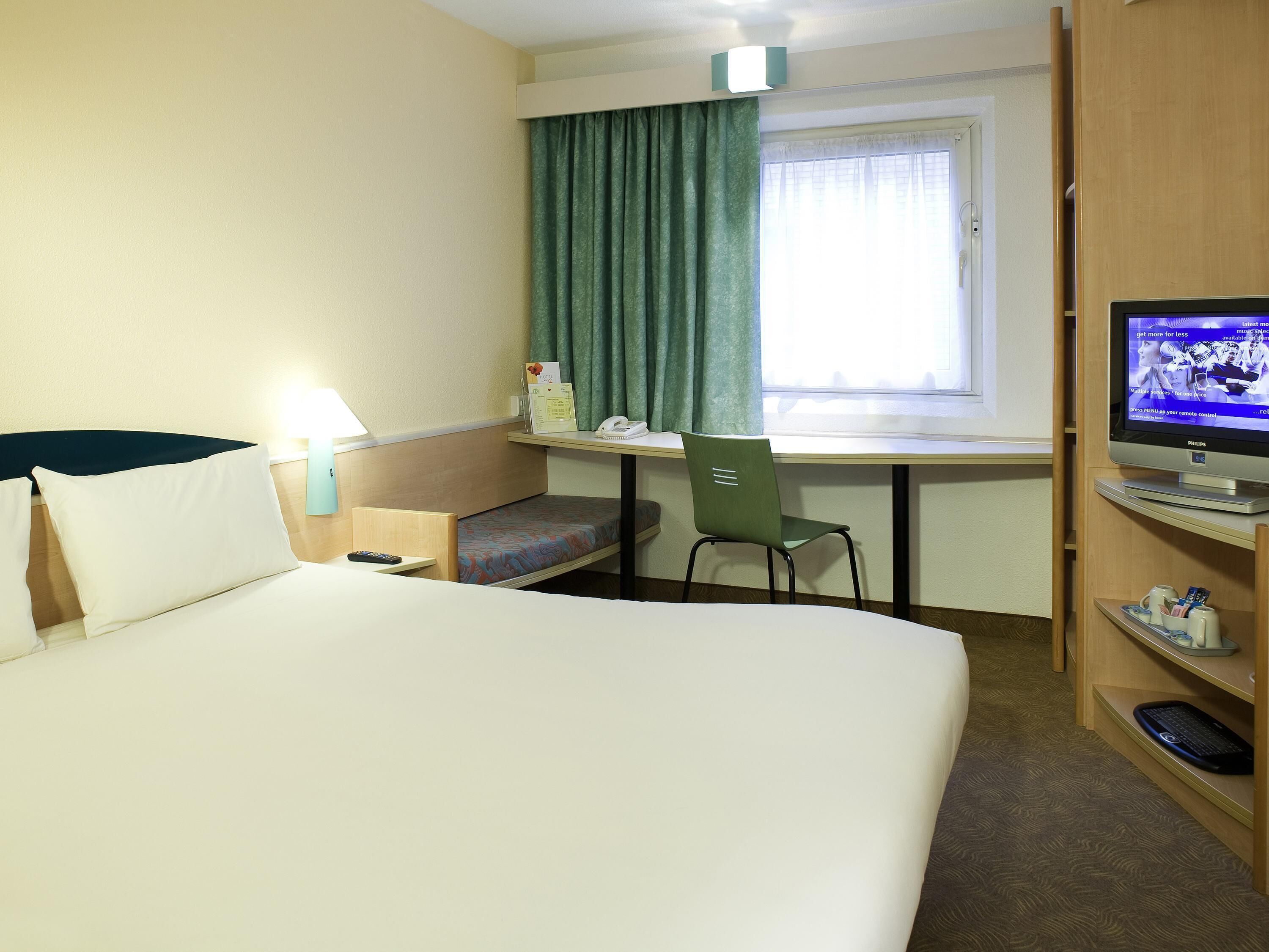 Habitación de hotel con cama, escritorio, silla, televisor y ventana con cortinas.