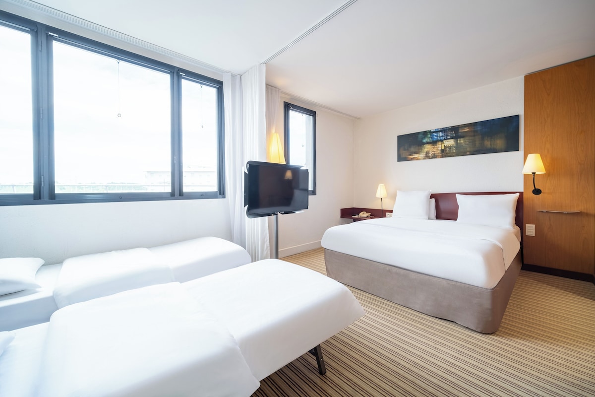 A hotel room with a bed, a TV, a bedside table with a lamp, and a painting on the wall.