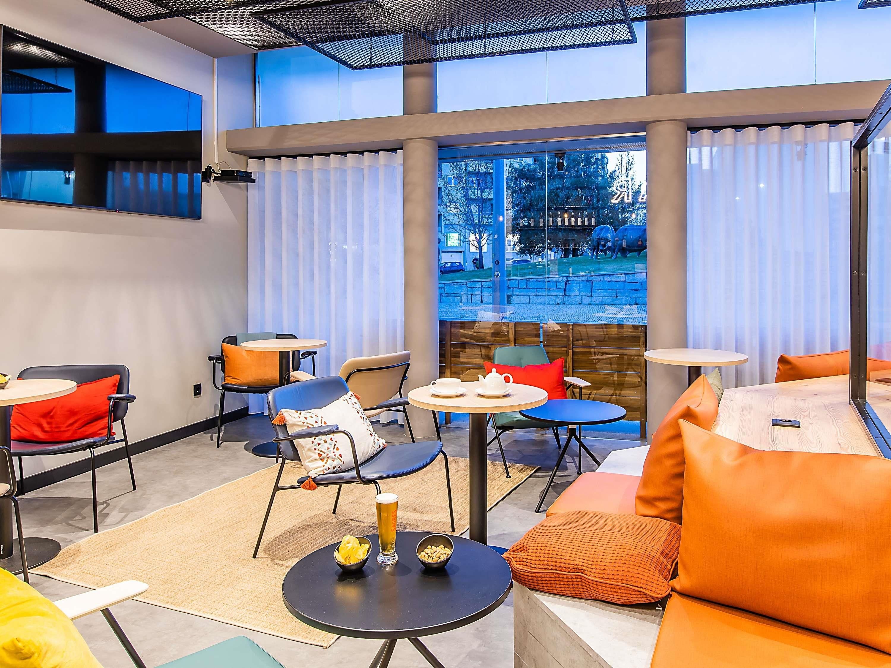 Un área de estar interior moderna con varias mesas y sillas, ventanas amplias con cortinas y vistas al exterior.
