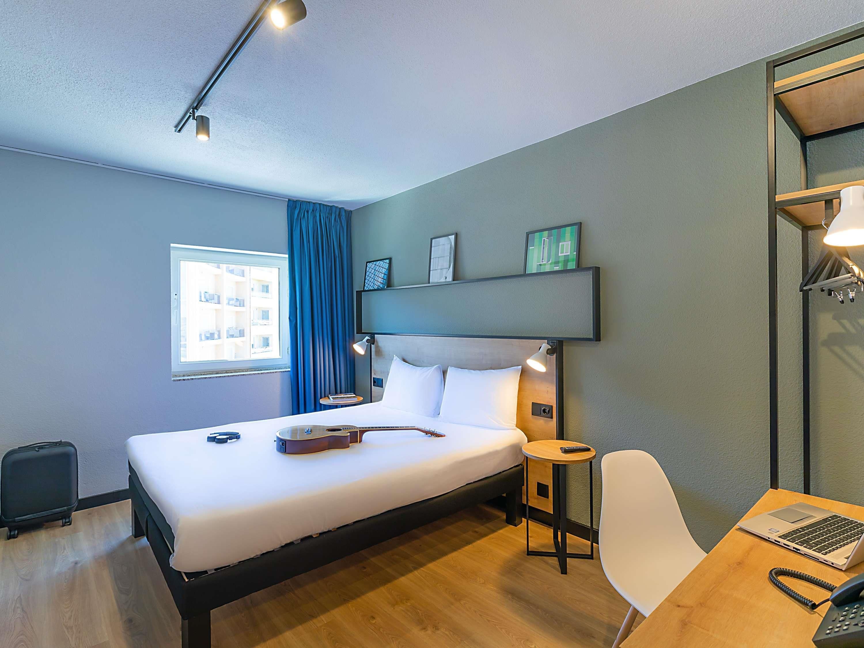 A hotel room with a bed, a desk with a laptop, a chair, a suitcase, and a window with curtains.