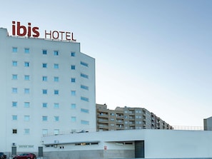 Ein Hotelgebäude mit einem Schild mit der Aufschrift 'ibis HOTEL' auf dem Dach, zahlreichen Fenstern und einem Parkplatz mit Autos.