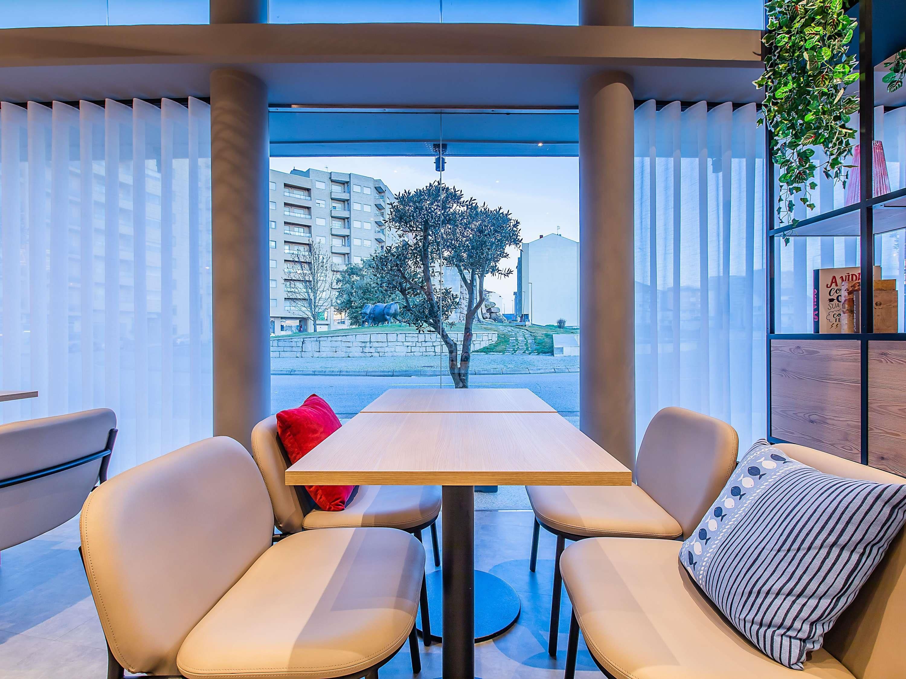 Un espacio interior moderno con asientos, una mesa y sillas, ventanas amplias con cortinas translúcidas y vistas a un paisaje urbano con edificios y un árbol.