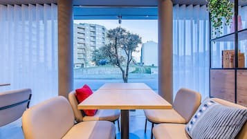 Un espacio interior moderno con asientos, una mesa y sillas, ventanas amplias con cortinas translúcidas y vistas a un paisaje urbano con edificios y un árbol.