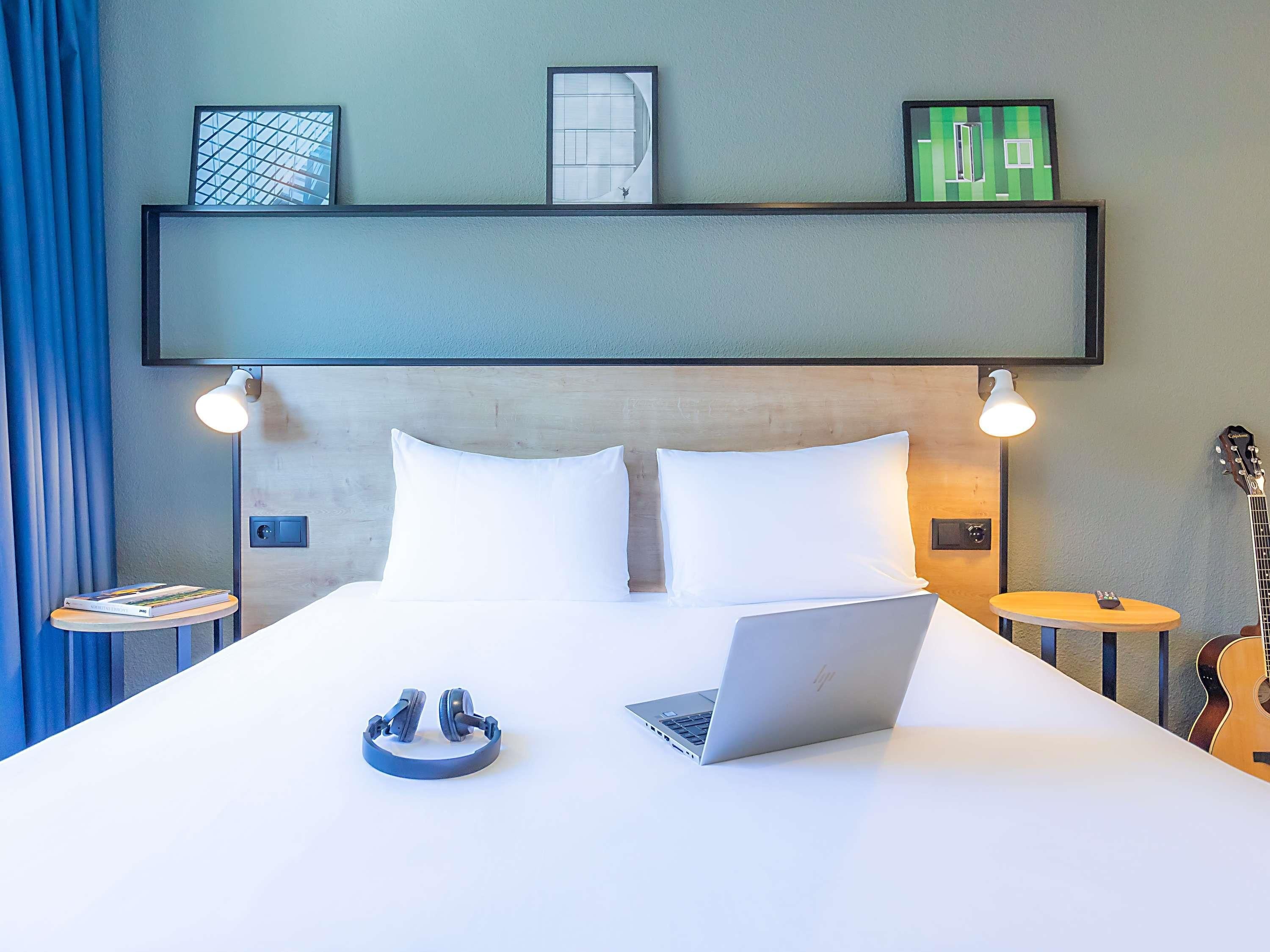 A neatly made bed with white pillows and a laptop on it. There are two bedside tables, one with a guitar and the other with a lamp.
