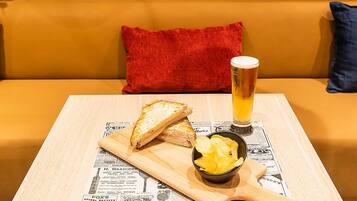 A wooden cutting board with a sandwich, a bowl of chips, and a glass of beer on a newspaper-covered table, placed on a yellow sofa with a red pillow.