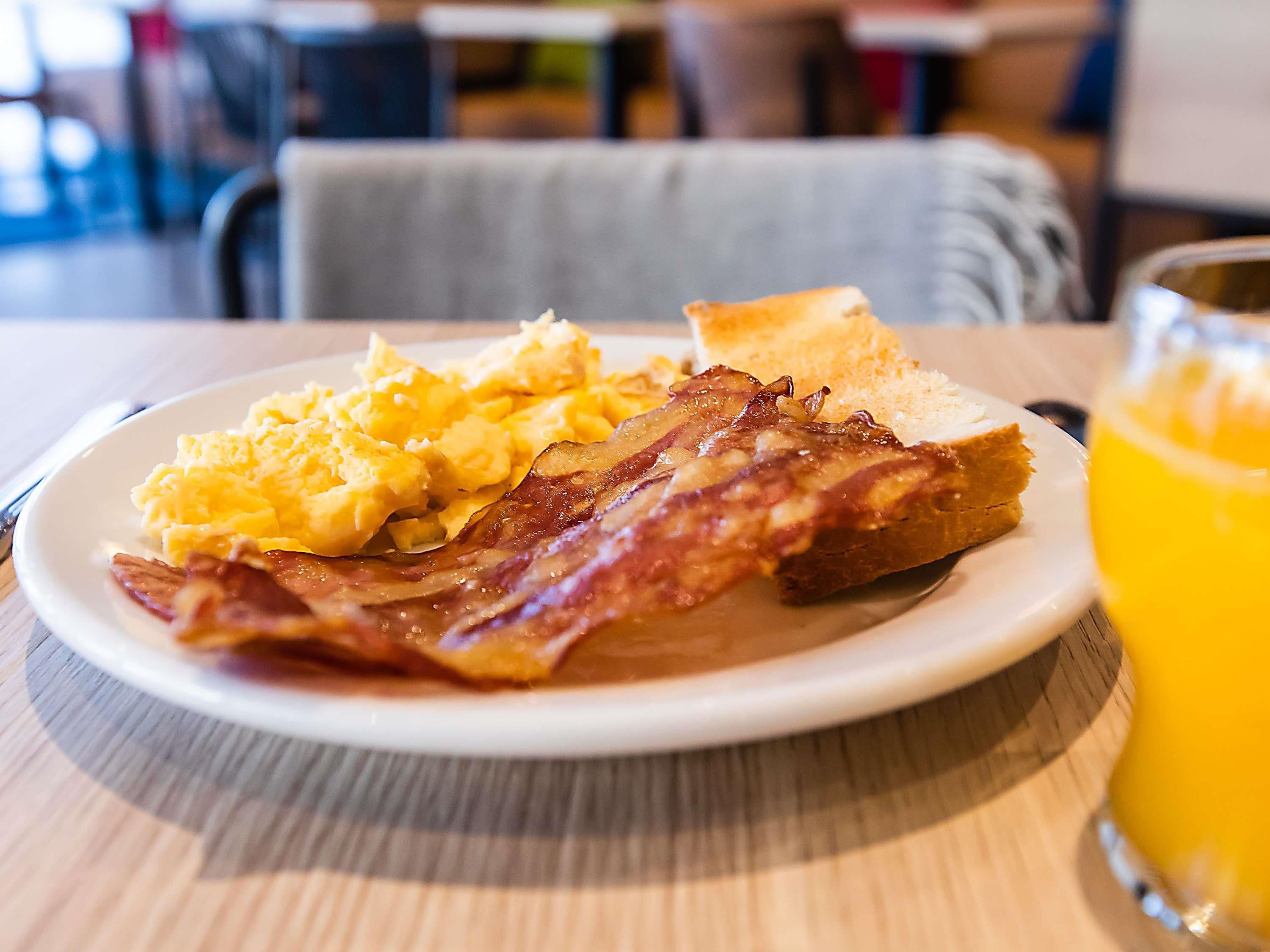 Un plato de desayuno con huevos revueltos, tocino, tostadas y un vaso de jugo de naranja sobre una mesa de madera.