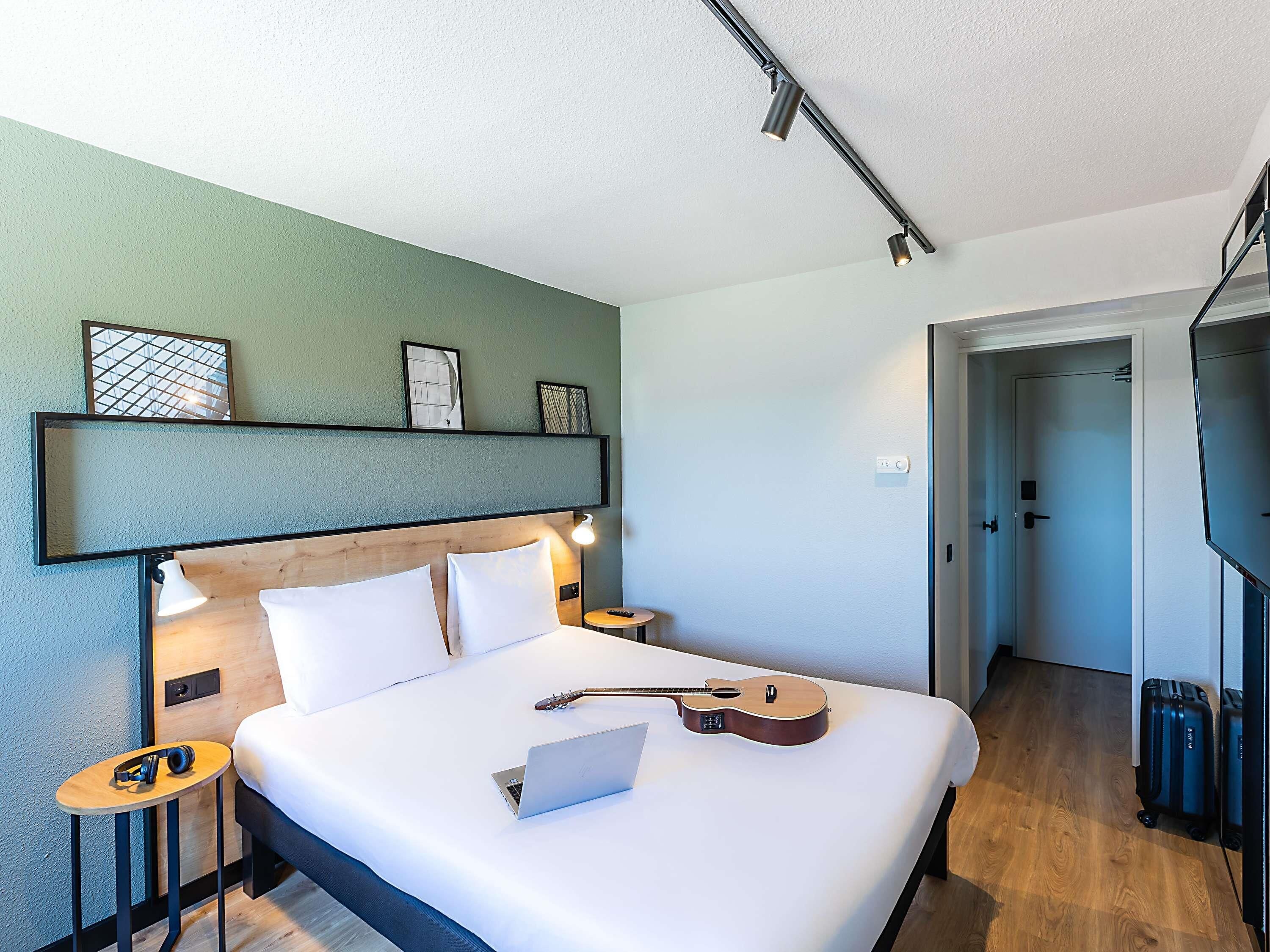 Ett hotellrum med en säng, en bärbar dator, en gitarr, en tv och en dörr som leder till ett annat rum.