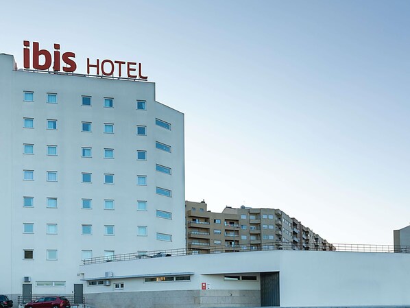 Un edificio de hotel con un letrero que dice 'ibis HOTEL' en la parte superior, múltiples ventanas y un estacionamiento con coches.