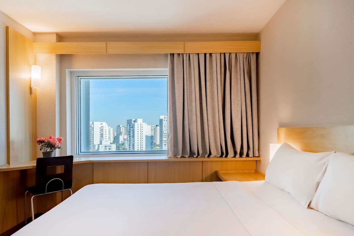 Hotelkamer met een groot bed, een bureau met stoel, een raam met gordijnen en uitzicht op stadsgebouwen.