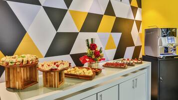 Uma mesa de buffet com diversos alimentos, uma máquina de café e um vaso decorativo com flores.