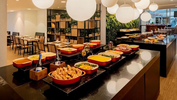 A buffet setup with various dishes and a wine bottle, surrounded by modern furniture and pendant lighting. - Ibis Sao Paulo Congonhas (São Paulo)