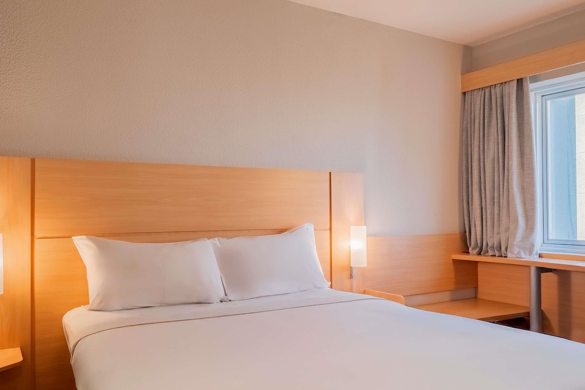 Een hotelkamer met een groot bed, een houten hoofdeinde, bedlampen en een raam met gordijnen.