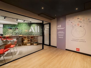 Een moderne hotellobby met een glazen wand, een groene wand met een logo, een zithoek met rode stoelen, en een wand met een wereldbol en tekst.