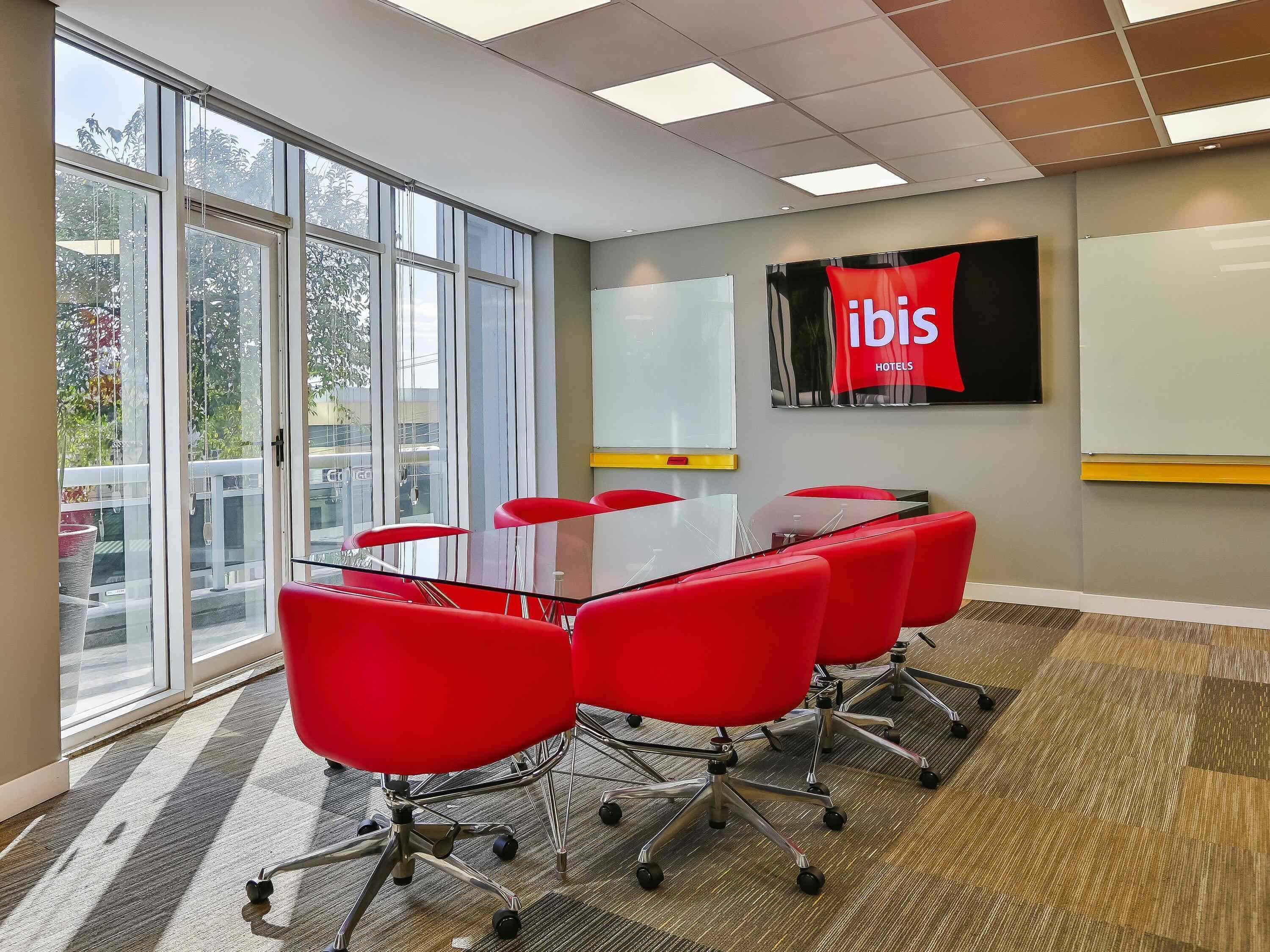 A conference room with a glass table, red chairs, a whiteboard, and a large window with a view of trees and buildings.