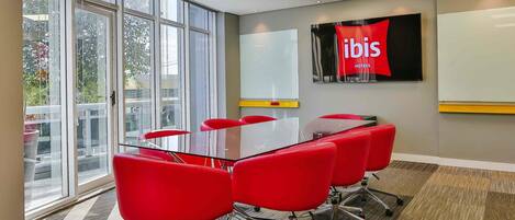 A conference room with a glass table, red chairs, a whiteboard, and a large window with a view of trees and buildings.