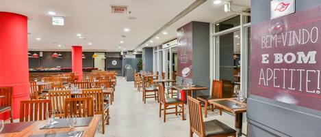 A spacious restaurant with multiple dining tables and chairs, a visible emergency exit sign, and a wall with a health and safety message.