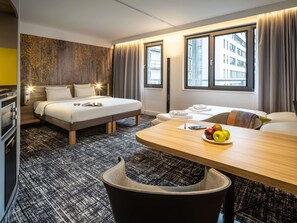 A hotel room with a bed, a desk with fruit, a chair, a table, and a view of buildings through the window.
