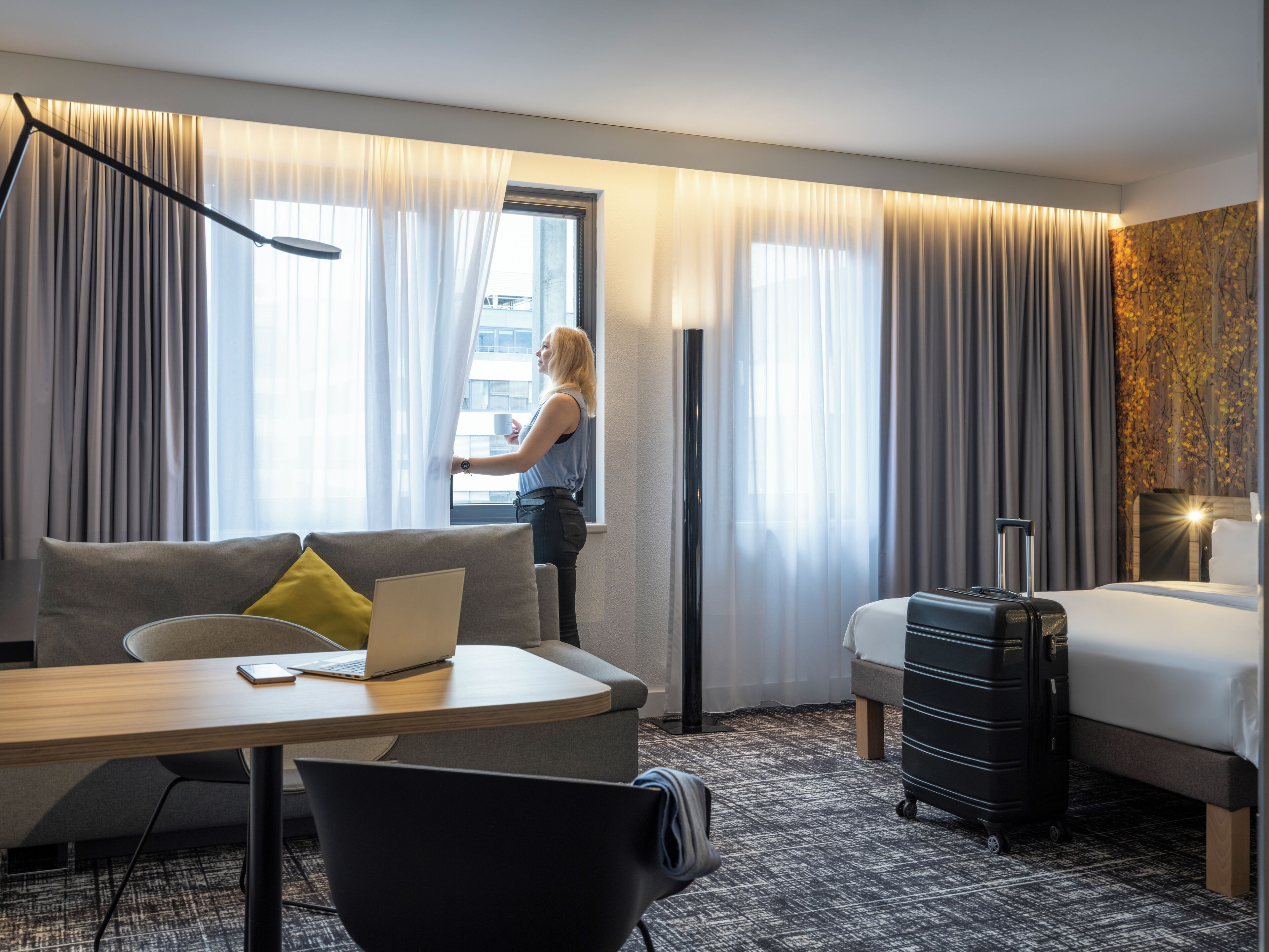 A modern hotel room with a sofa, a desk with a laptop, a bed, and a suitcase.