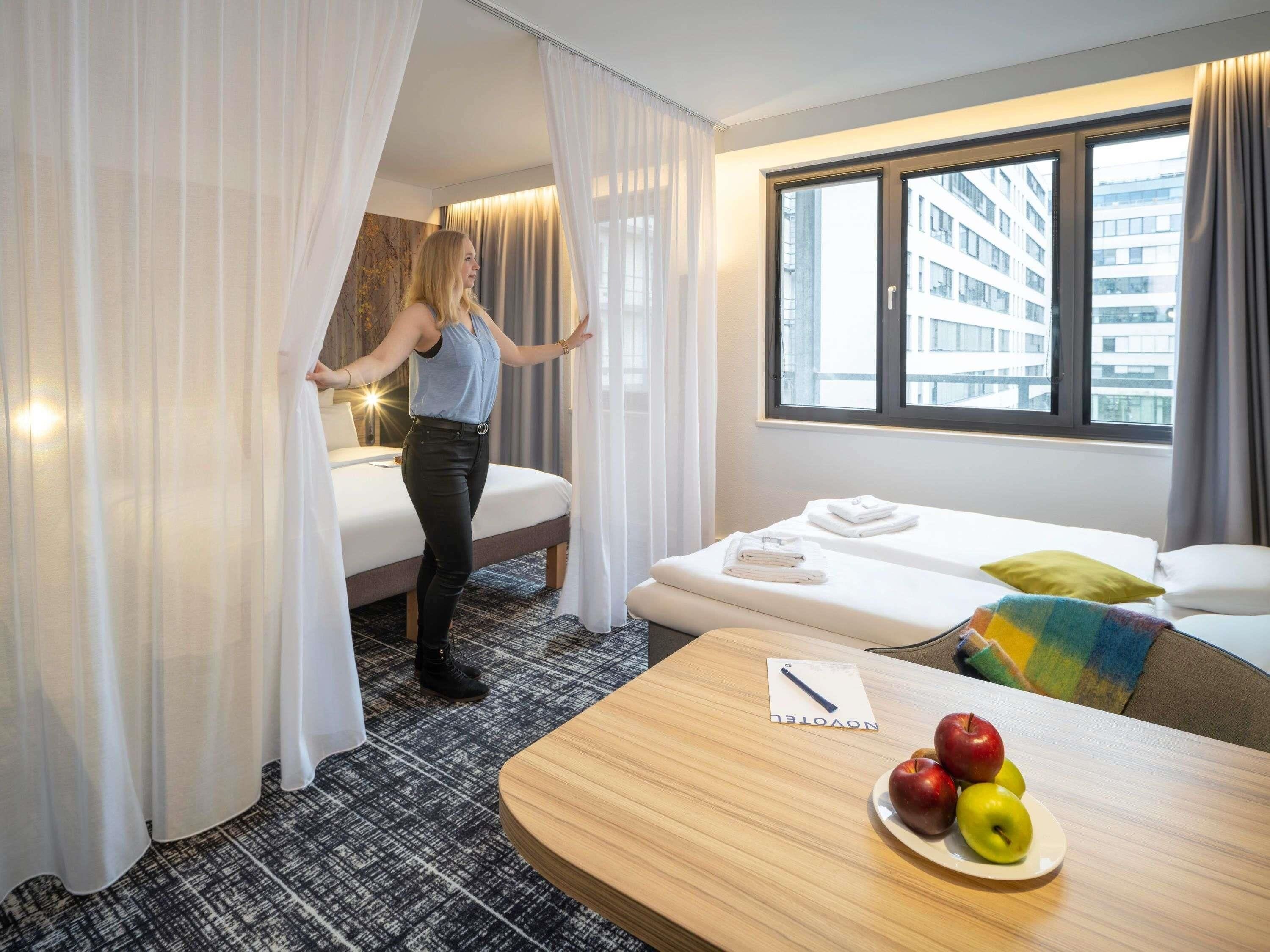 A hotel room with a bed, a small table with apples, a window with a view of buildings, and a curtain partially drawn.