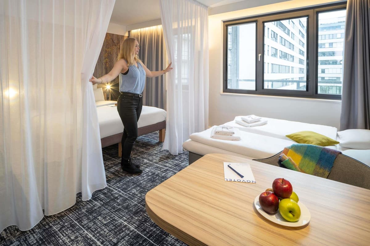 A hotel room with a bed, a small table with apples, a window with a view of buildings, and a curtain partially drawn.