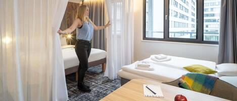 Habitación de hotel con cama, una mesita con manzanas, ventana que ofrece vistas a edificios y cortina entreabierta.