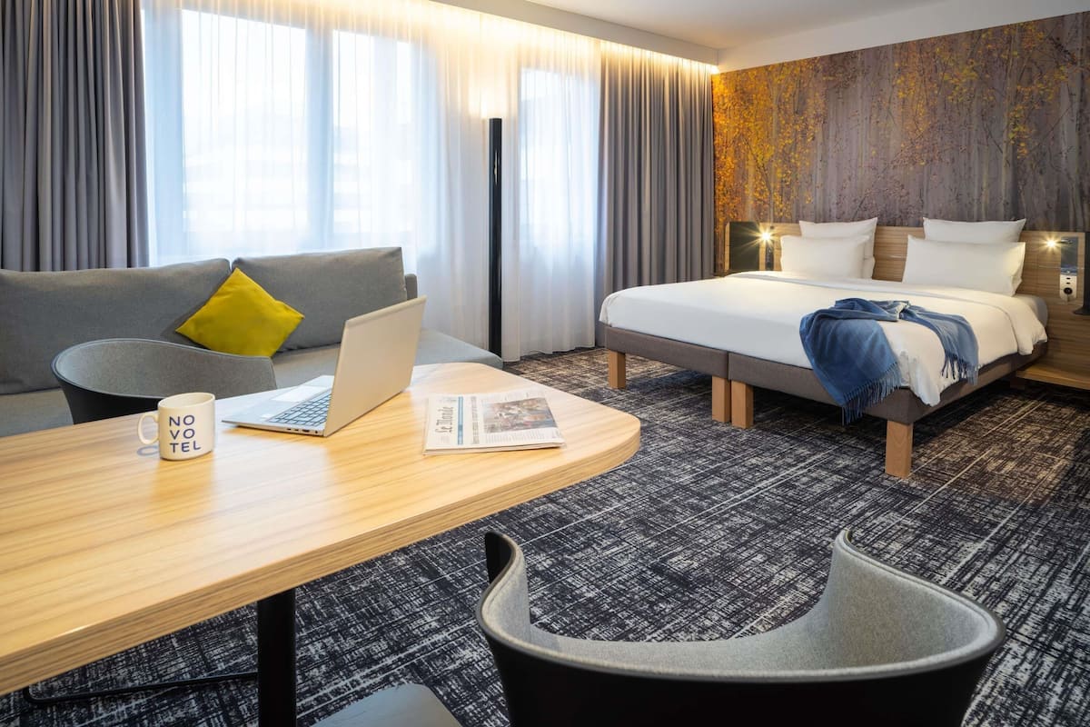 Quarto de hotel moderno com um sofá cinzento, uma escrivaninha de madeira com um laptop e um jornal, uma cama com um cobertor azul e uma janela grande com cortinas.