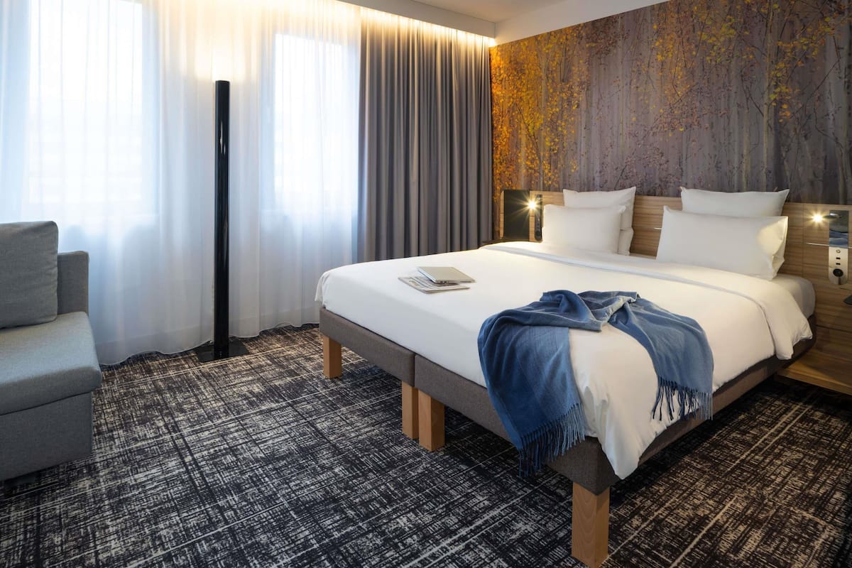 Quarto de hotel com uma cama grande, uma escrivaninha, uma cadeira e vista para árvores pela janela.