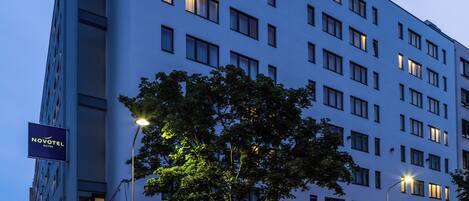 Un hotel moderno con un llamativo letrero de 'Novotel', una entrada bordeada de árboles y un paso de peatones al frente.
