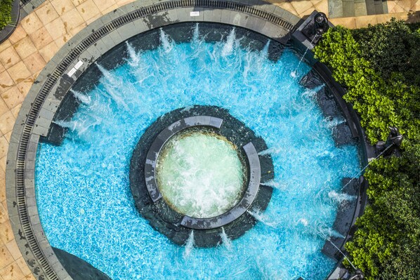 A circular pool with a central fountain, surrounded by a paved walkway and greenery.