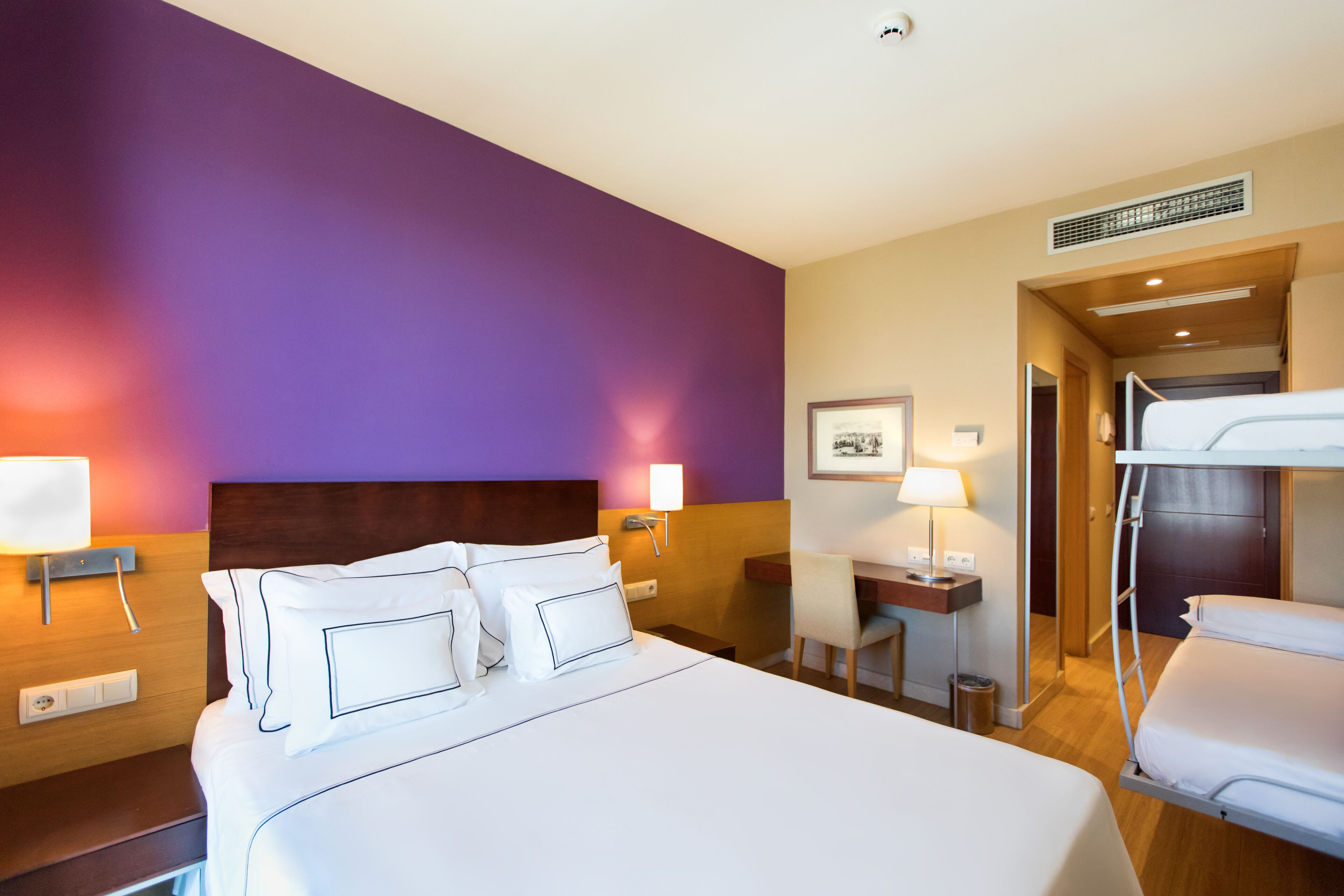 A hotel room with a large bed, bedside tables, a desk, and a bunk bed.