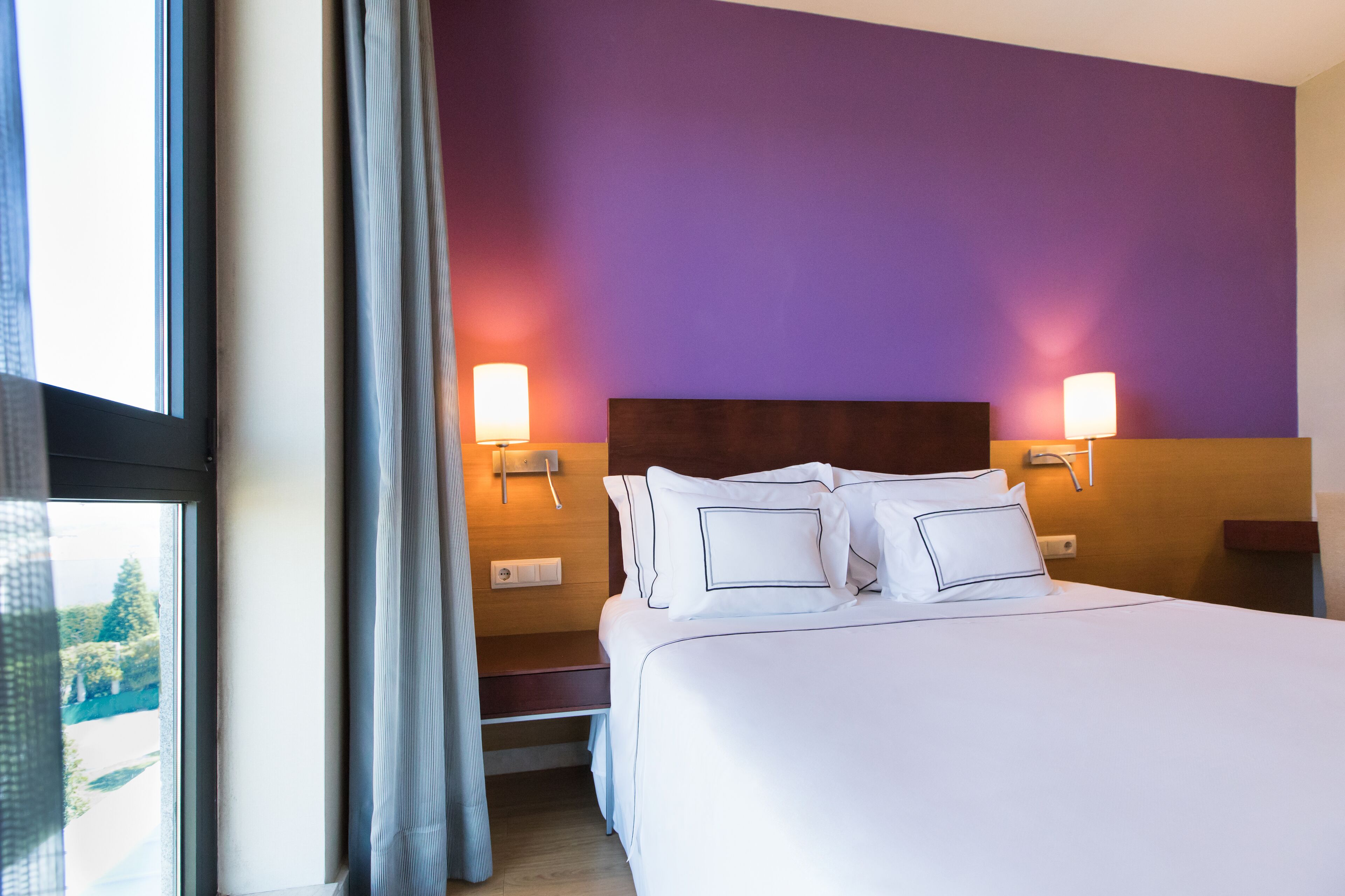 Habitación de hotel con una cama grande, dos lámparas de noche, cabecera de madera y vistas a zonas verdes.