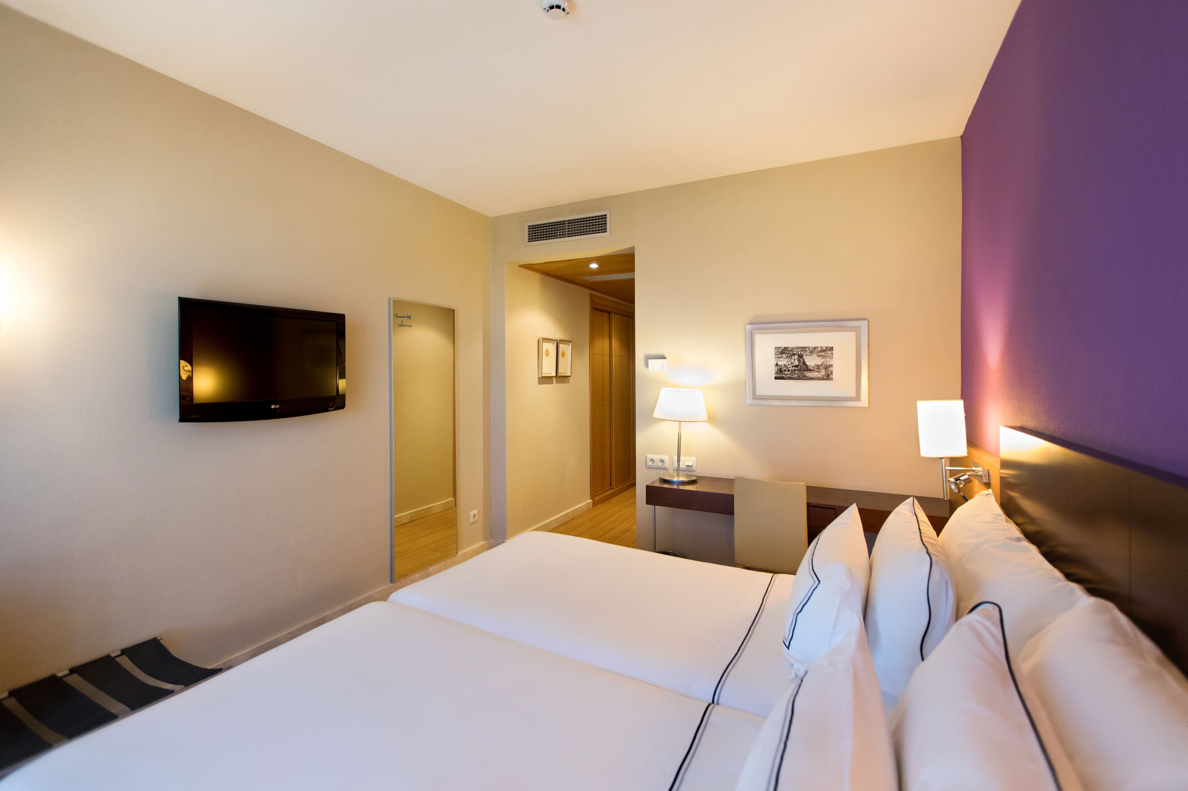 A hotel room with a bed, a television mounted on the wall, a desk with a lamp, and a framed picture on the wall.