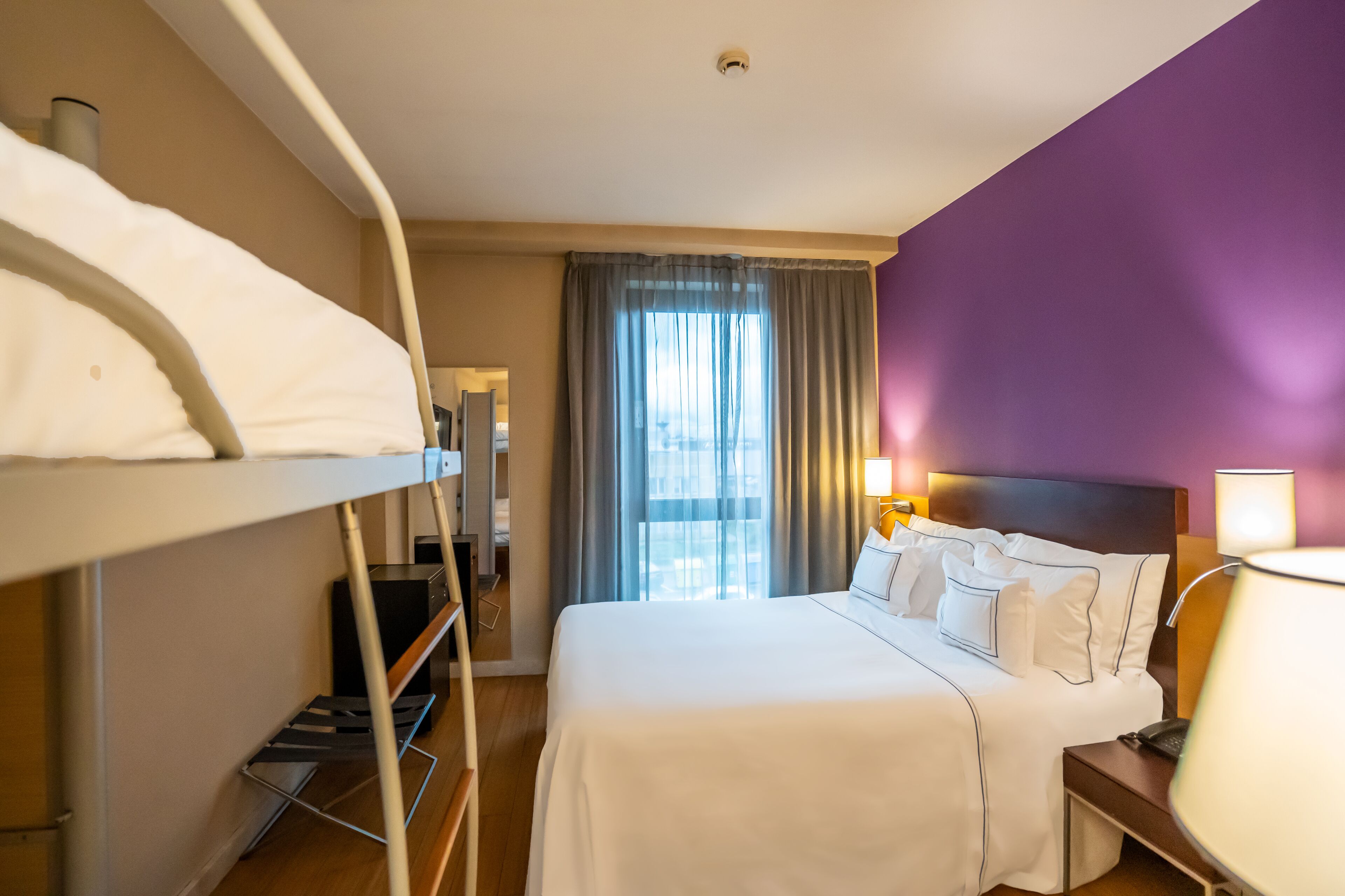 A hotel room with a bunk bed, a single bed, a desk with a lamp, a window with curtains, and a view of the city.