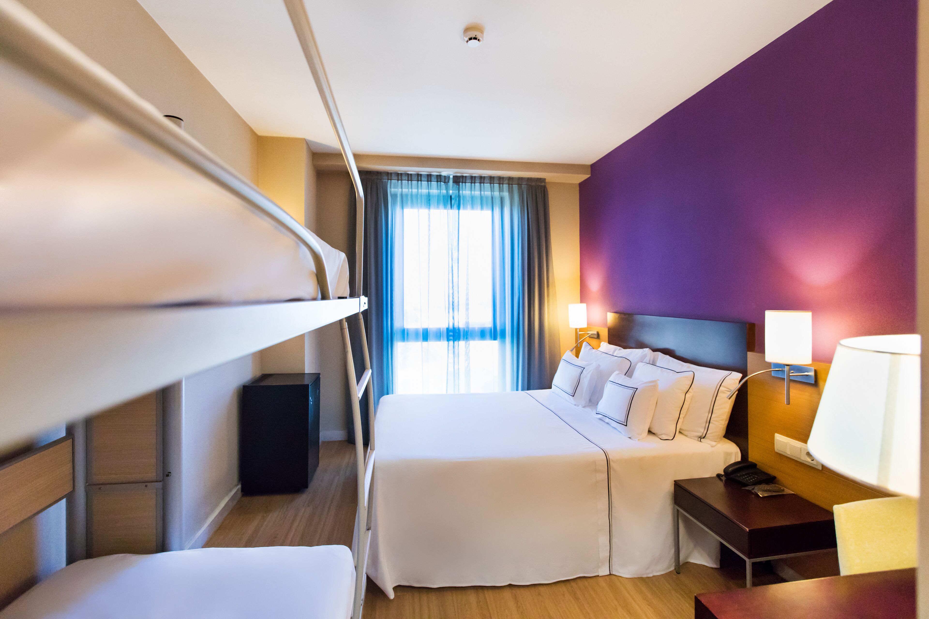 Een hotelkamer met een stapelbed, een nachtkastje met een lamp, een bureau en een raam met gordijnen.