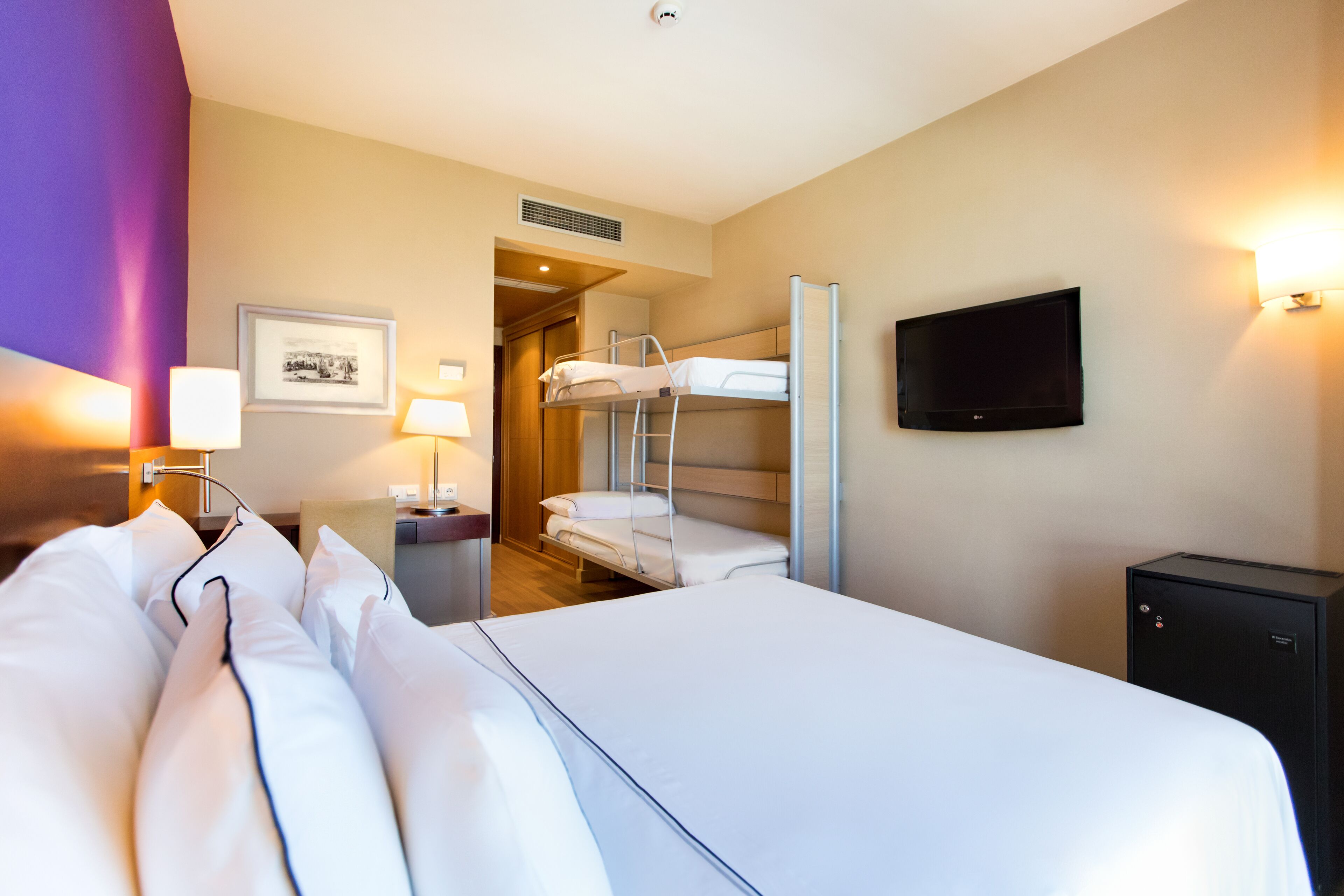 Habitación de hotel con una cama grande, una cama litera, un escritorio con computadora, un televisor y un cuadro en la pared.