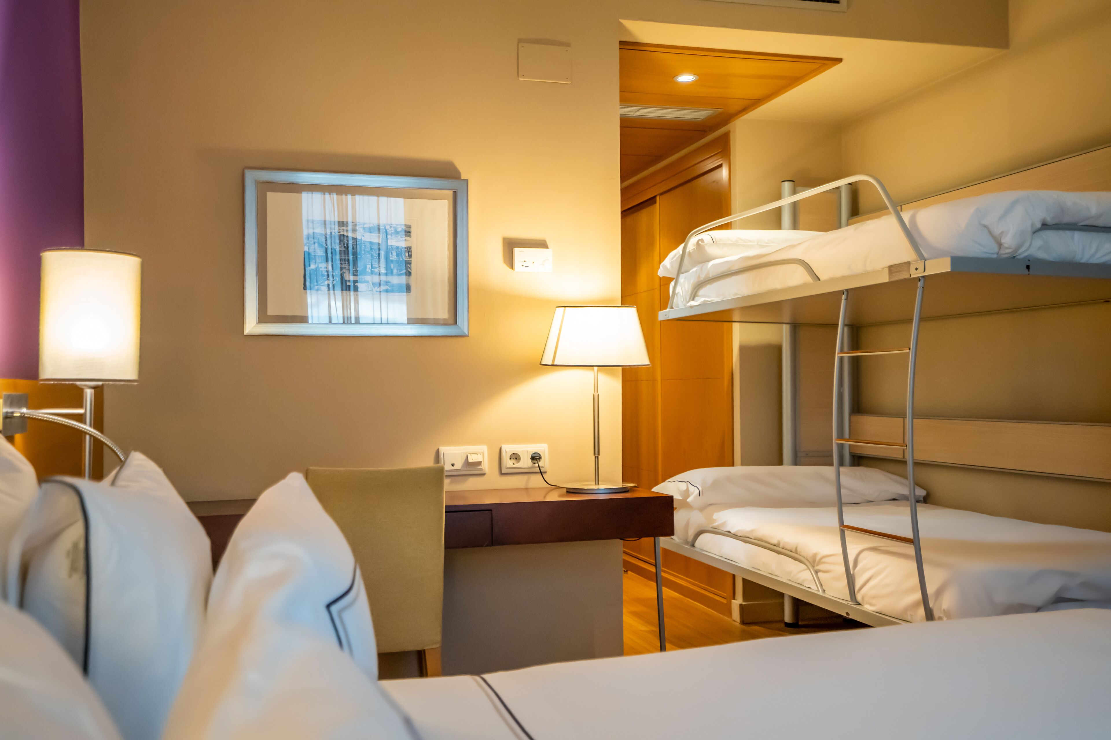 A hotel room with bunk beds, a desk, a chair, a lamp, and a framed picture on the wall.