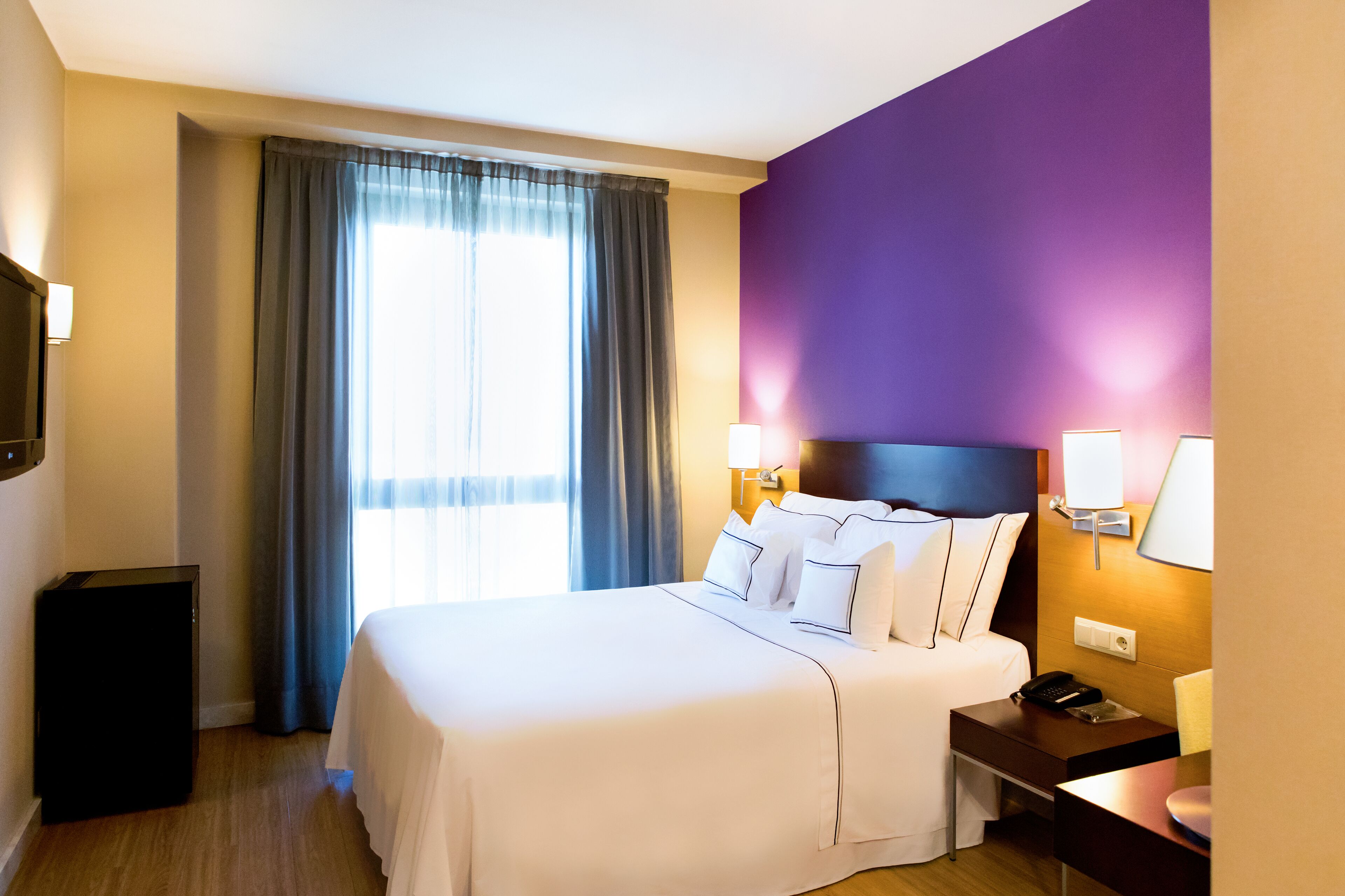 A hotel room with a large bed, a desk, a chair, a TV, and a window with curtains.