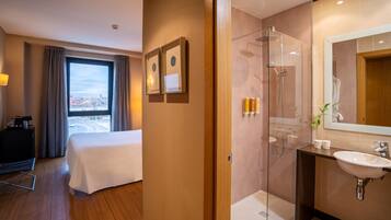 Una habitación de hotel con cama, un ventanal con cortinas, un baño con ducha de vidrio y un lavamanos.