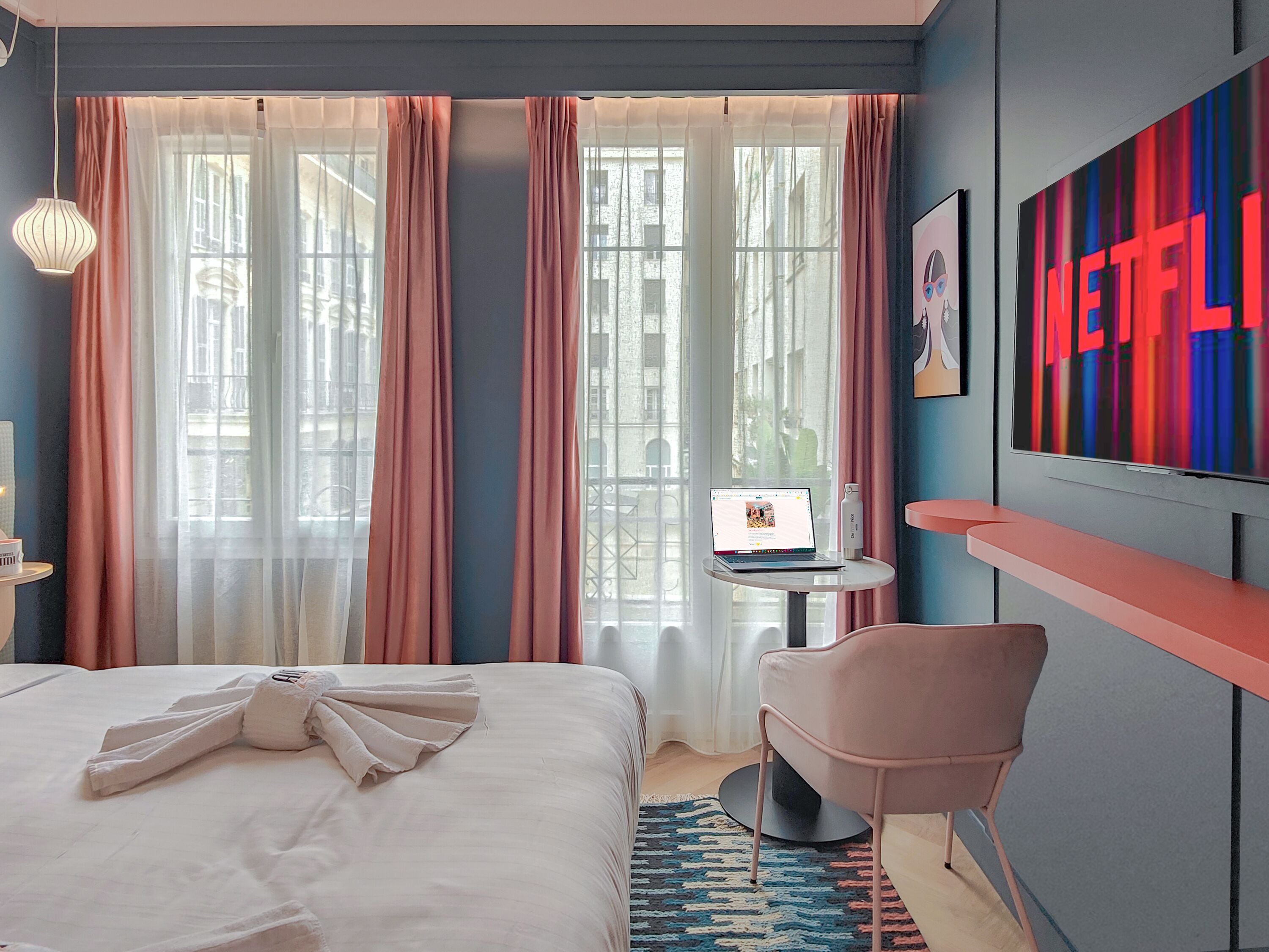 A hotel room with a bed, a desk with a laptop, a chair, a TV with a Netflix logo, and a window with curtains.