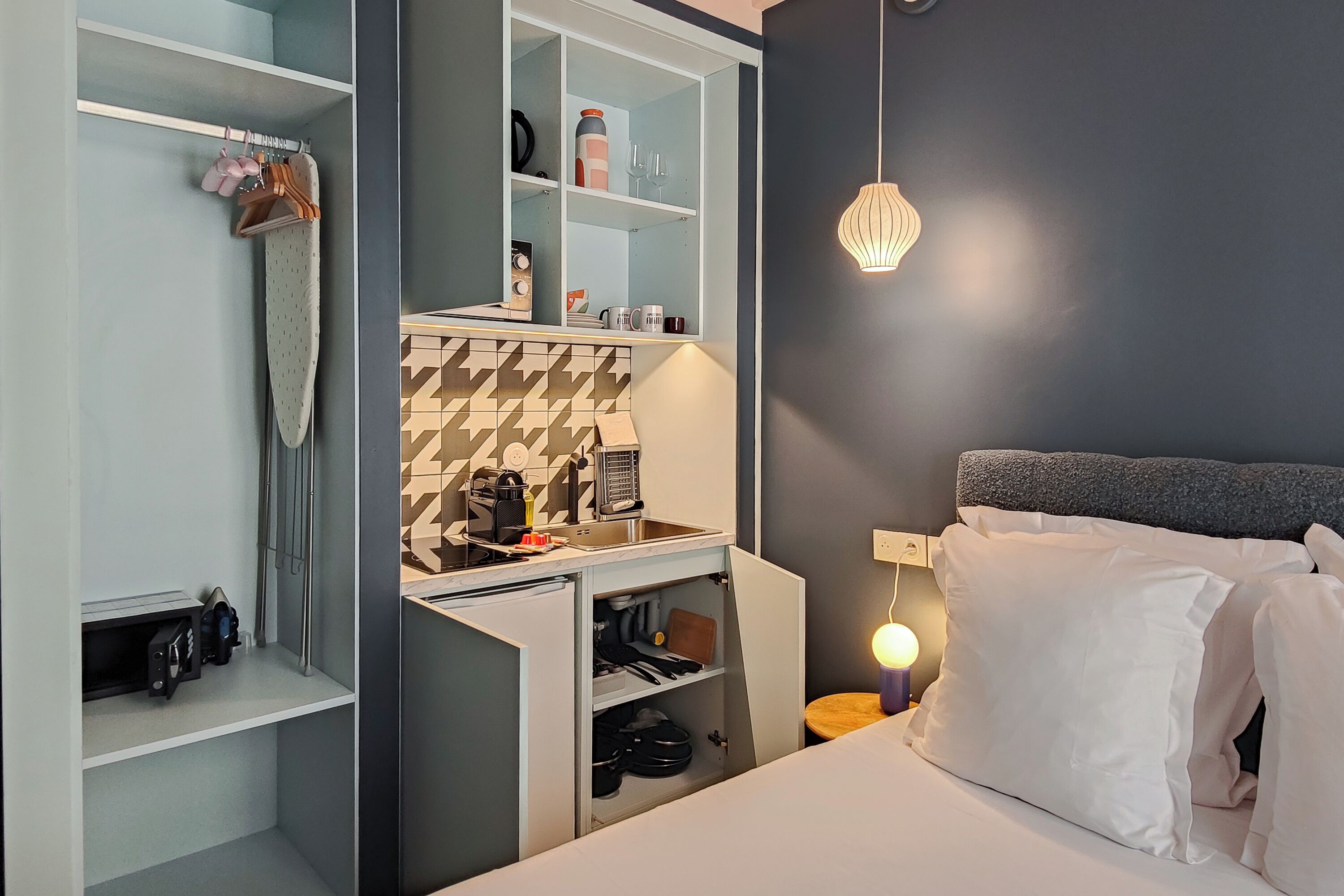 A compact hotel room with a bed, a wardrobe, a small kitchenette, and a bedside table with a lamp.