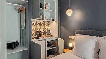 A compact hotel room with a bed, a wardrobe, a small kitchenette, and a bedside table with a lamp.