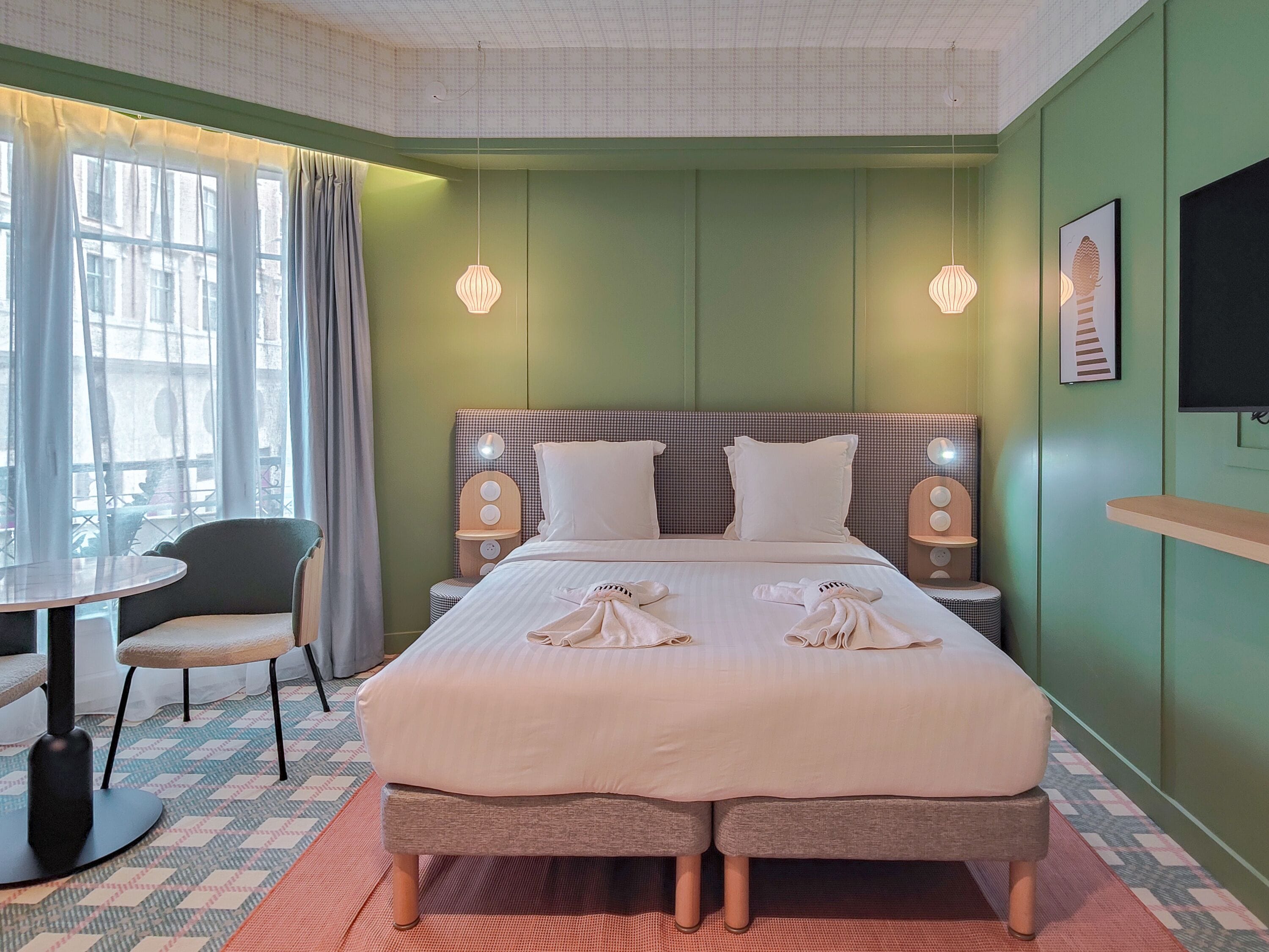 A hotel room with a large bed, two bedside tables, a chair, a small round table, and a window with curtains.