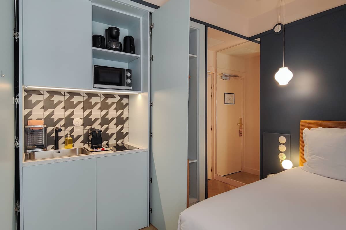Habitación de hotel compacta con kitchenette, una cama con cabecera y una mesita de noche con lámpara.