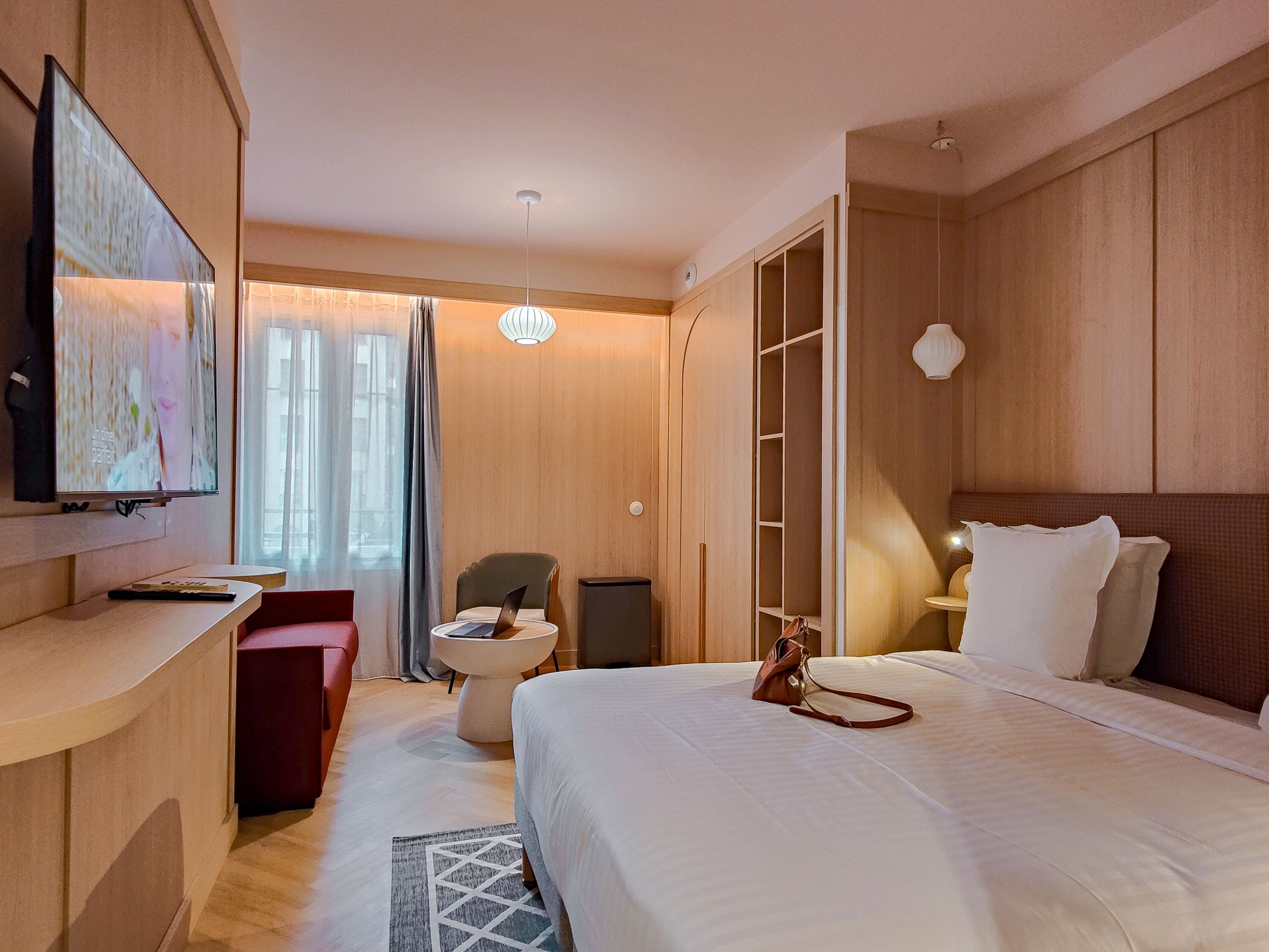 A hotel room with a bed, a desk with a laptop, a TV, a wardrobe, and a seating area.