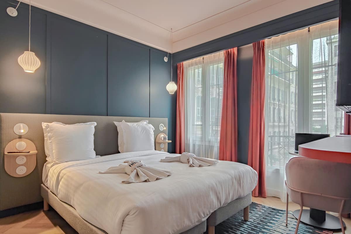 Una habitación de hotel moderna con una cama grande, mesitas de noche, un escritorio y vistas al exterior a través de amplios ventanales.