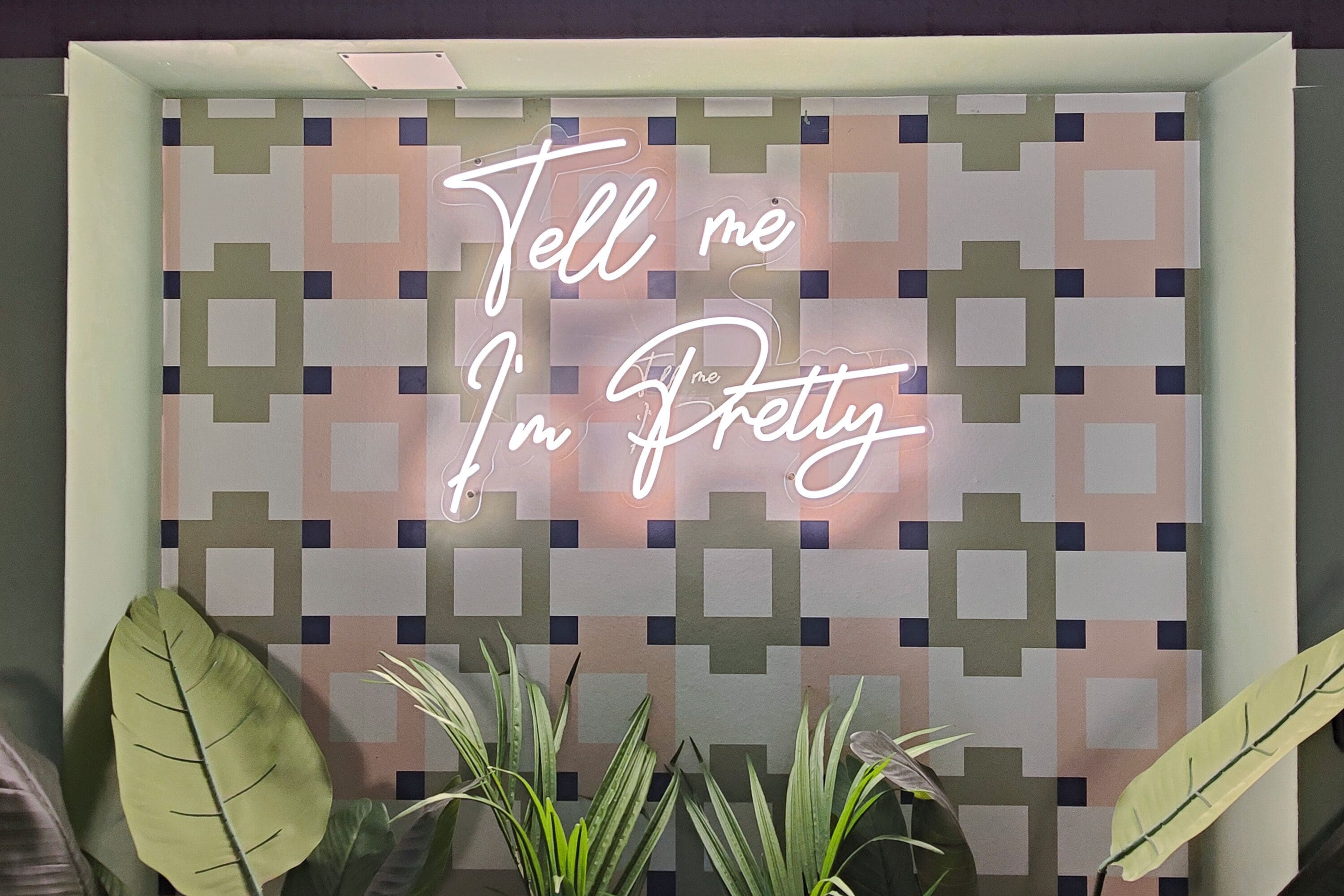 Ein Neon-Schild mit dem Schriftzug 'Tell me I'm Pretty' vor einer Wand mit geometrischen Mustern.