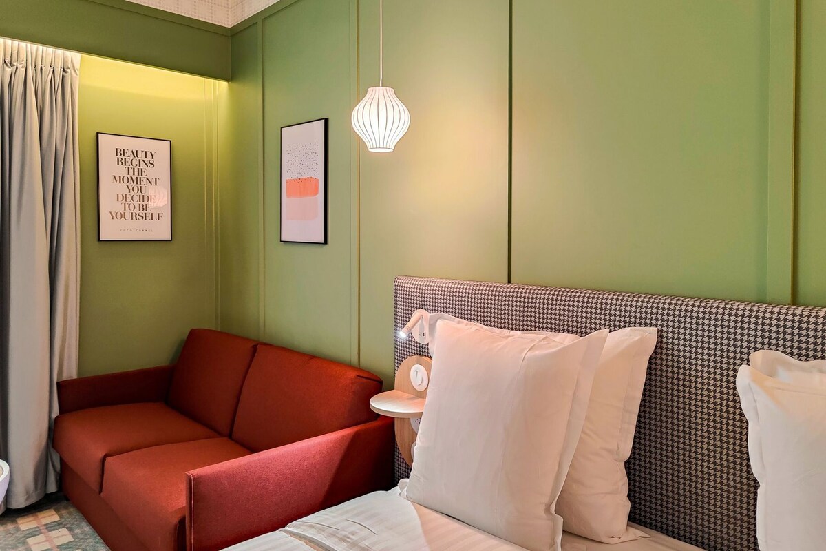 A room with a bed, a red sofa, a small table, and a hanging light fixture.