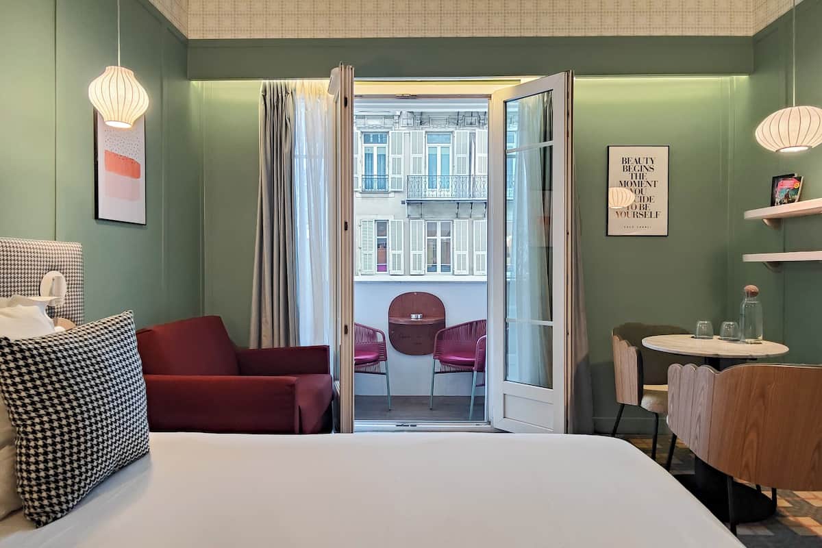 Habitación de hotel con una cama, un sillón rojo, una mesita con vasos, una silla y vistas a un balcón exterior.